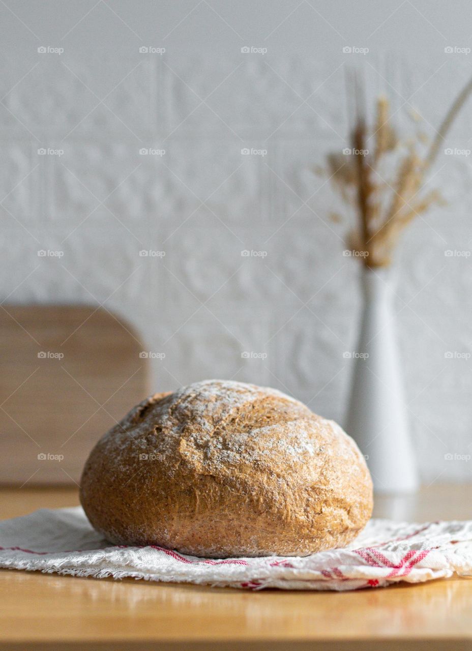 One round wheat bread with cereals on a kitchen napkin lies on a wooden table against a blurred white brick wall with a split board and a dried flower vase, close-up view. The concept of baking bread.