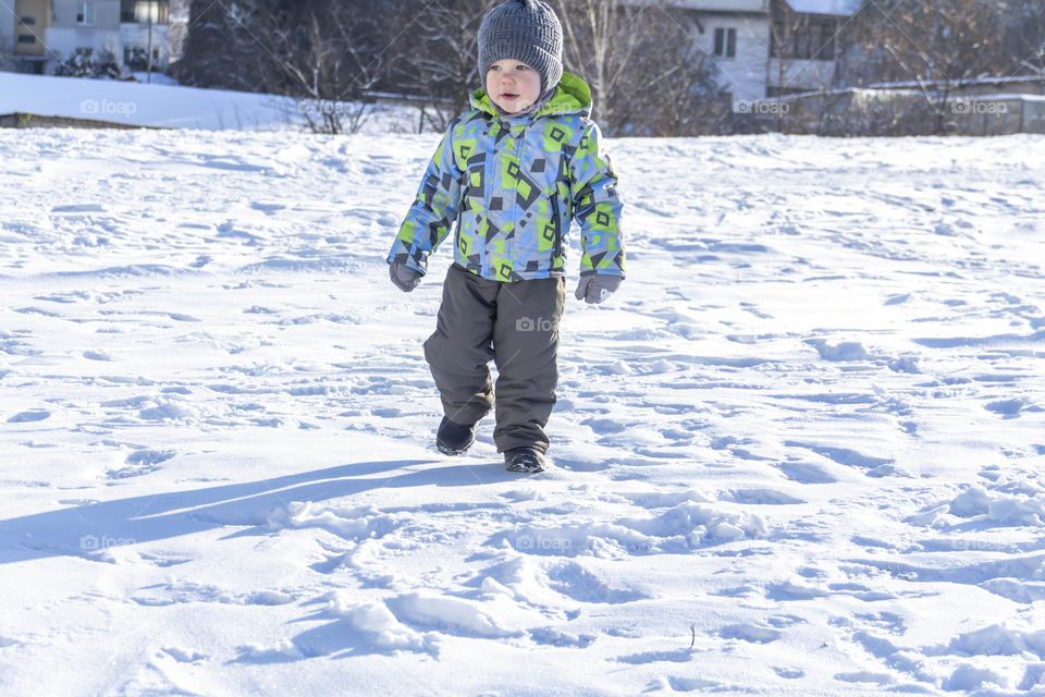A child with a serious expression on his face in winter clothes jackets, pants, hat and boots in winter on the white snow on the street and in the park in nature plays winter fun.