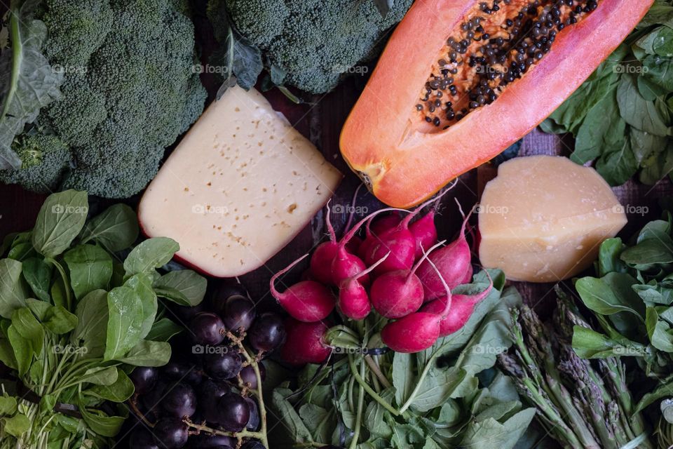 A wooden table full of vegetables, herbs, berries, fruits and cheese. Flat lay with cut papaya, broсcoli, arugula, tomatoes, mango, cilantro, radish, cheese, Parmesan