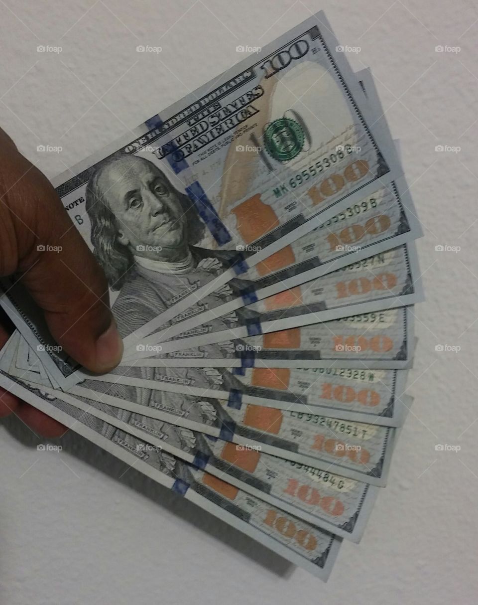 a man's hand holding eight $100 bills fanned out against the wall, ready to be spanked.