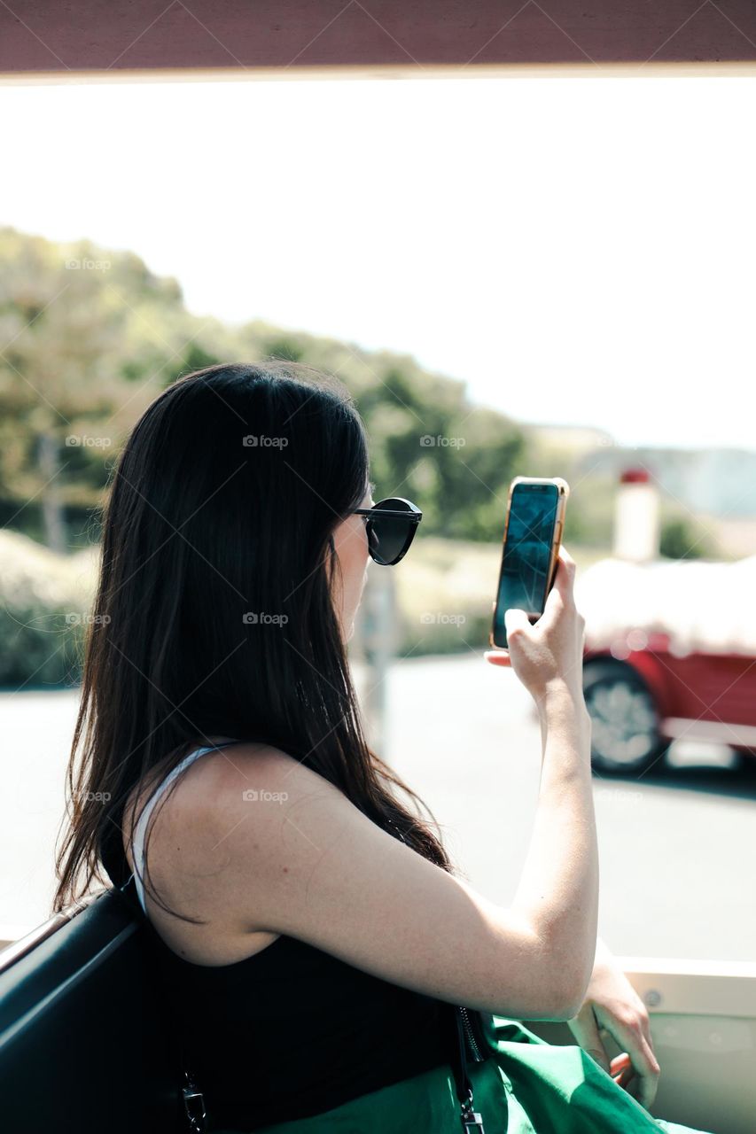 Beautiful young brunette girl with long hair in a black t-shirt shoots a landscape on a mobile phone while sitting in a tourist train in Normandy France, close-up side view.Concept using tesnology.