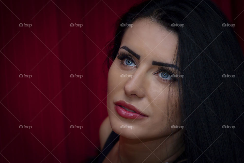 a portrait of a woman looking upward in front of a curtain. she has red lips and blue eyes.