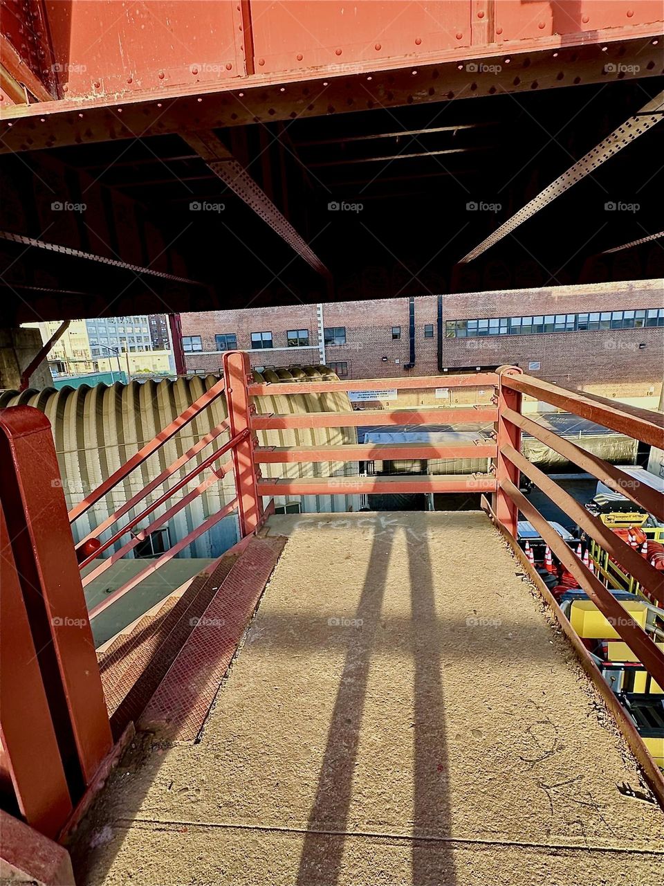 This is the red metal staircase of the “Pulaski Bridge” at “Newtown Creek” in LIC, Queens. The afternoon sun casts elongated shadows onto the paved middle platform complimentary to the strong geometric shapes of the bridge. 2024. Hypnotic Productions