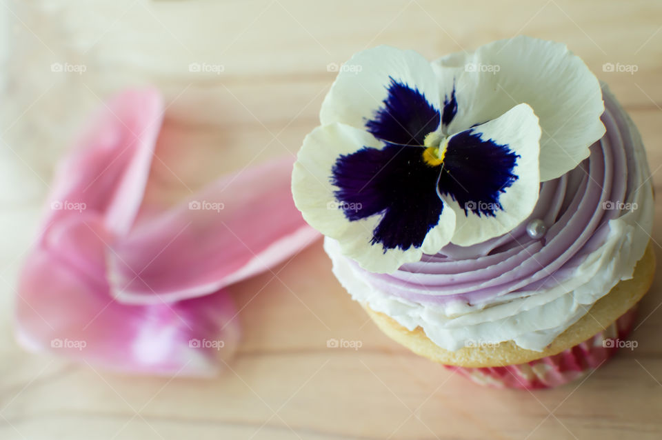 Delicate cup cake with lavender and vanilla buttercream frosting and edible pansy flower decoration on wood background with fresh pink petals 