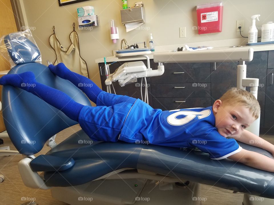 Blonde boy, in blue soccer uniform, plays across dentist chair, smiling.