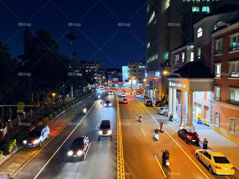 A busy street at night in Changhua, Taiwan. Several scooters and cars drive by. To the right are buildings, and to the left are trees.