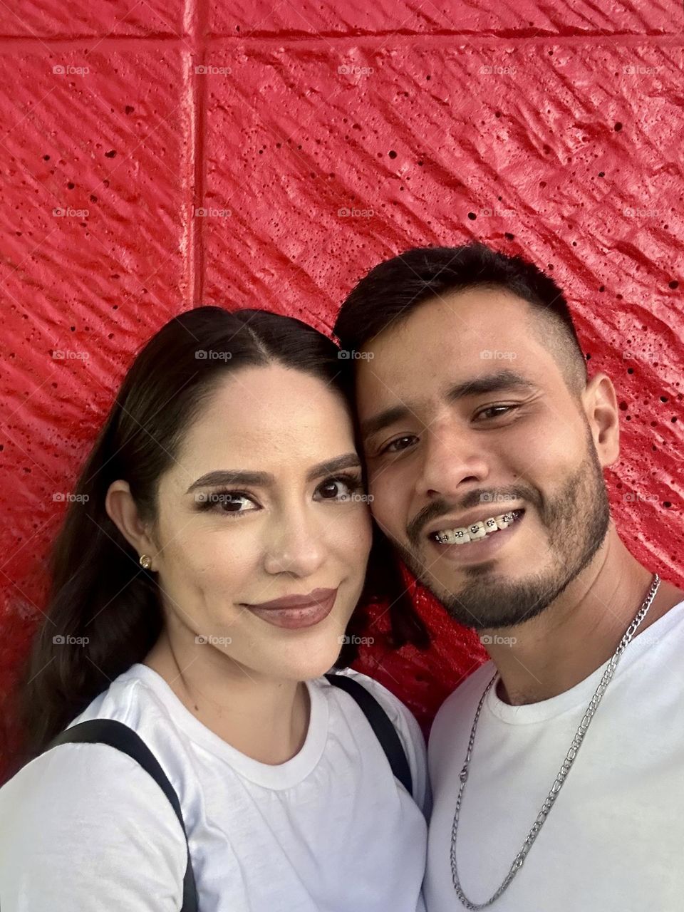 Young people taking a selfie, in a red wall at the background while they smiling.