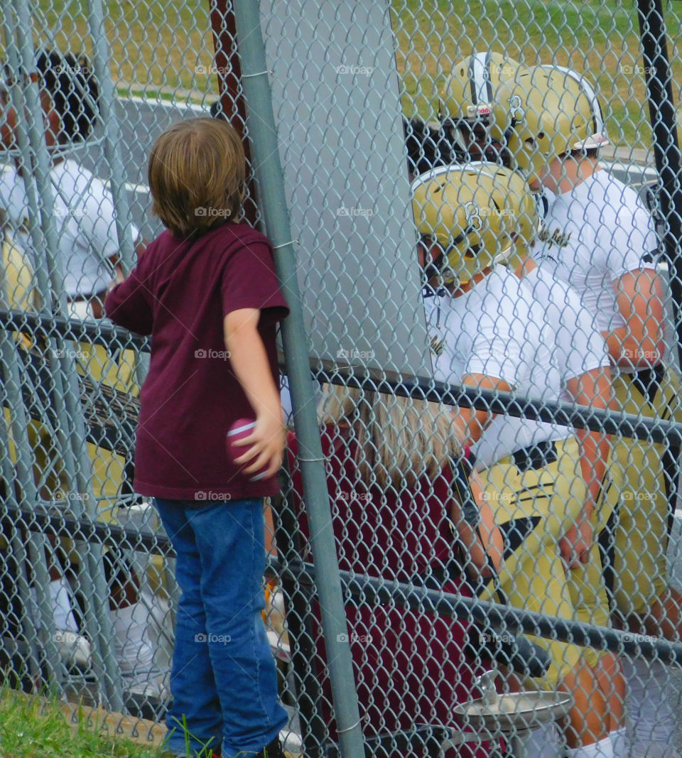 Can I play? Does anybody hear me? Can I play? Young boy holding a small football stands at the fence during a football game wanting to be noticed!