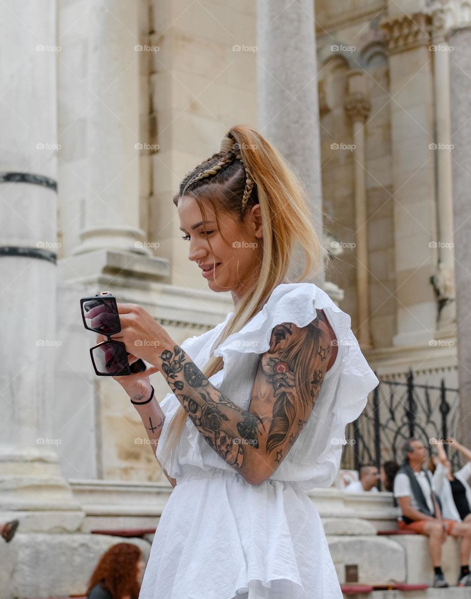Beautiful woman with tattoo on her arm wearing white dress, using mobile phone in street in city