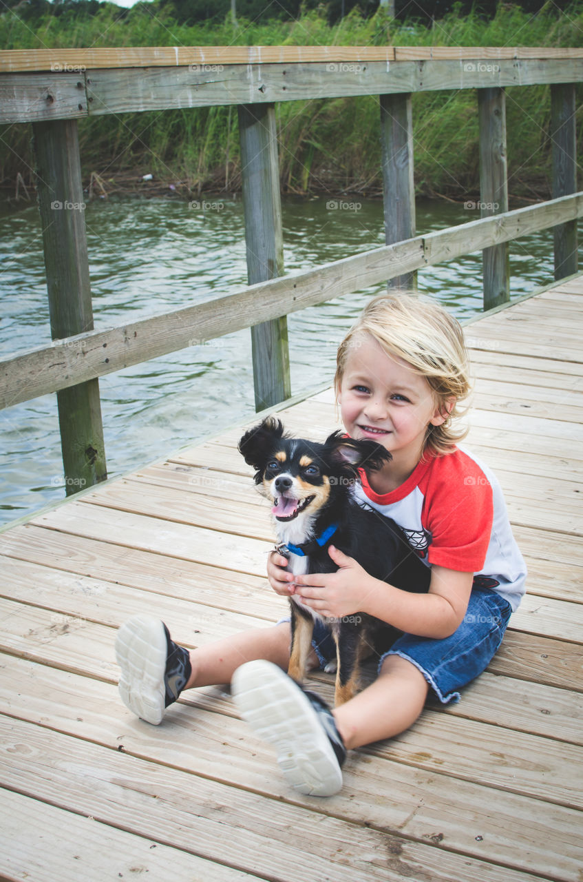Little boy with his pet dog is sitting on pier