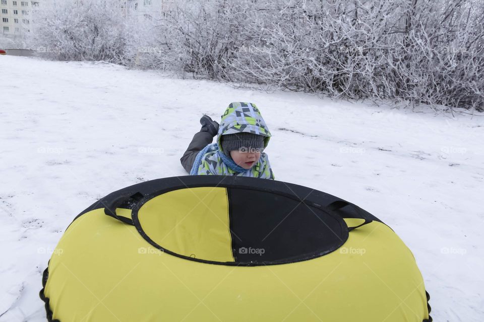 A small, carefree boy walks in the white snow in winter and rides a tubing in the park, near trees in the snow.