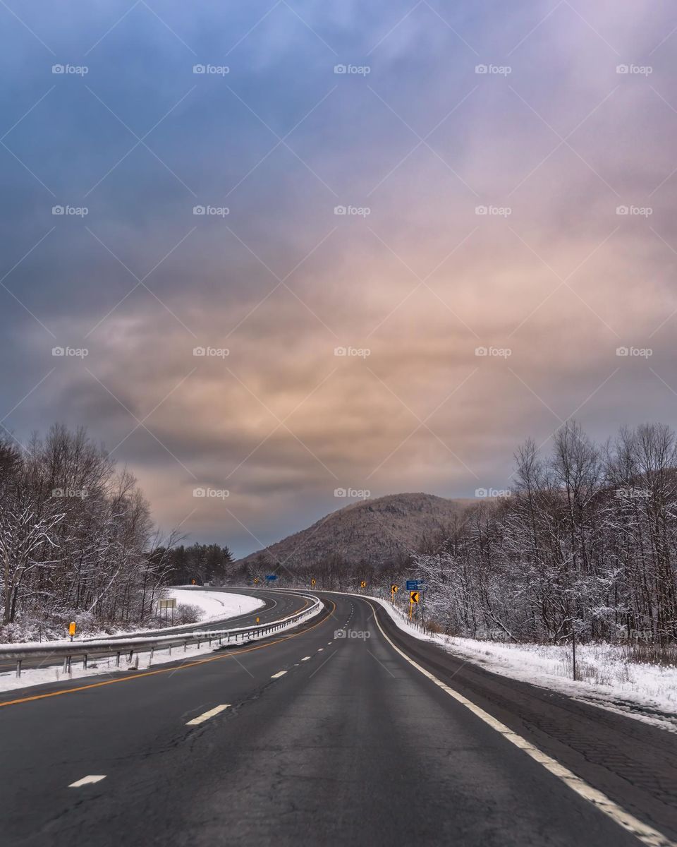 Dramatic storm clouds at sunset over a long winding road through the mountains, with snow covering the landscape and trees