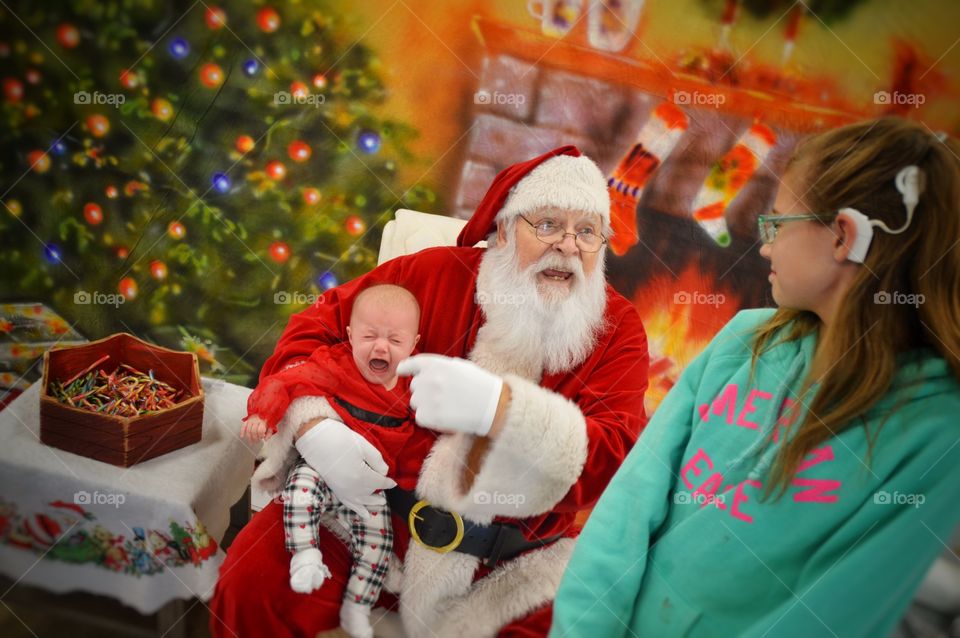 Funny photo of an unhappy baby on Santa's lap. Sister has to laugh as Santa tries to calm the little one.