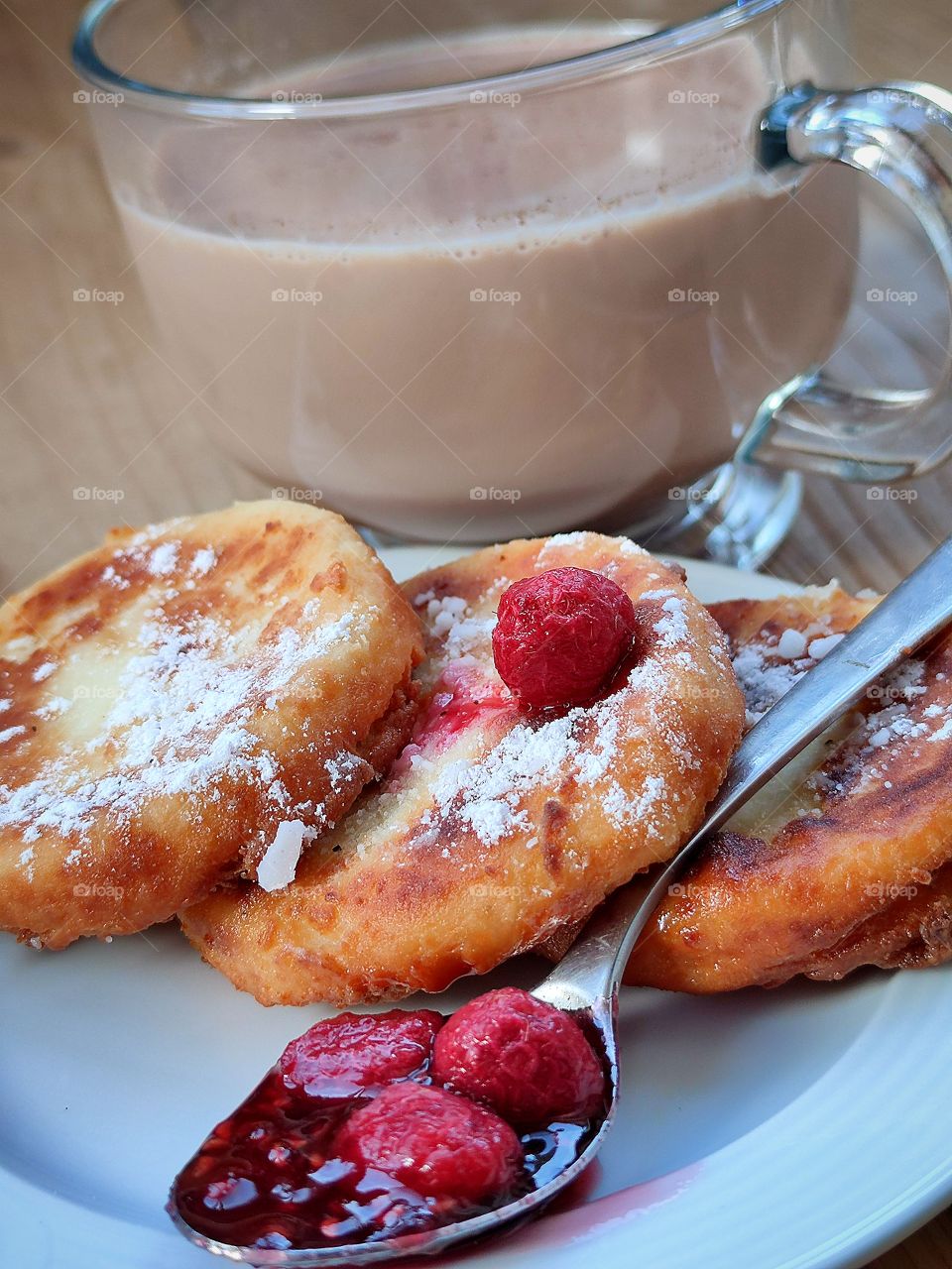 Breakfast. On the plate. three cheesecakes, sprinkled with powdered sugar. On the plate is a spoon with raspberry jam and fresh raspberries. There is a cup of hot cocoa next to the plate