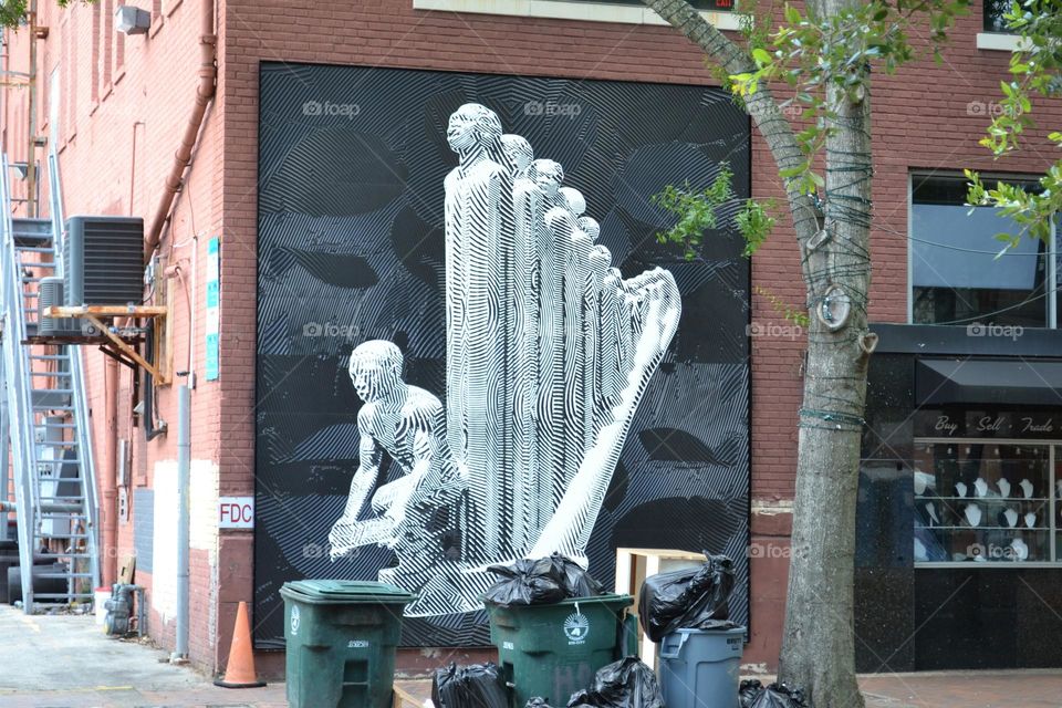 A black and white three dimensional mural of a man kneeling and a harp made of men on a city wall in front of green garbage cans and garbage bags next to a r tree