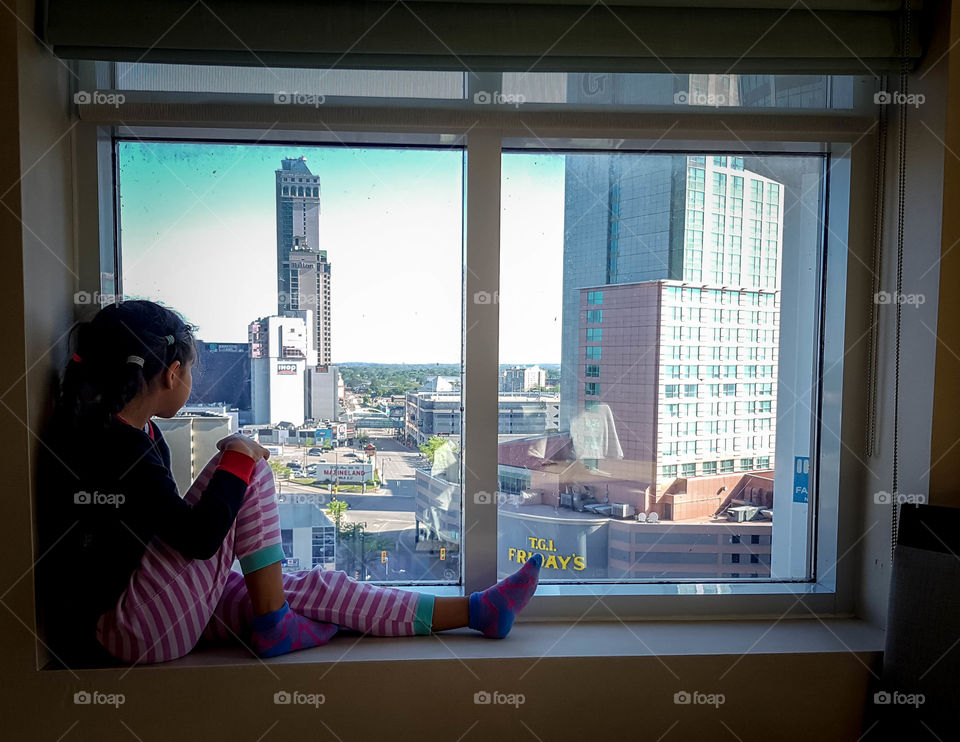 Little girl is sitting on a window sill looking at a cityscape below