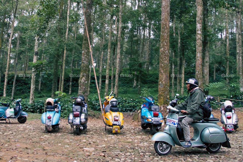 Portrait of an Asian man riding a VBB Classic Vespa in a national park in the city of Bogor with a row of vespas parked in front of green trees in the background.