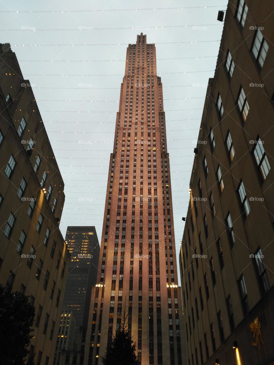 View from below of the Rockefeller building appreciating its large size, side of buildings with rectangular windows in which the light from their surroundings is reflected and Christmas lights hanging between them.