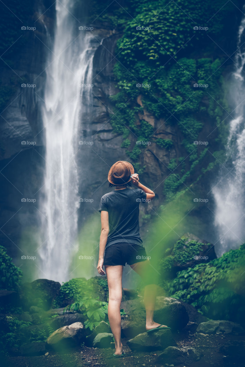 Girl in a straw hat stands on the background of a waterfall. Bali island.
