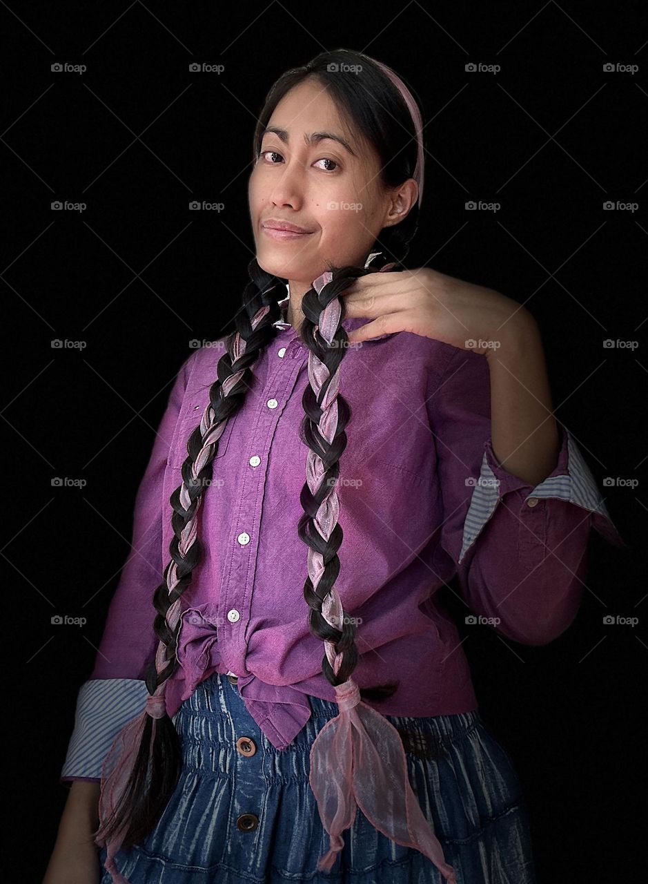 Portrait of a woman with braided long haired wearing purple long sleeved shirt and a blue skirt against black background. 