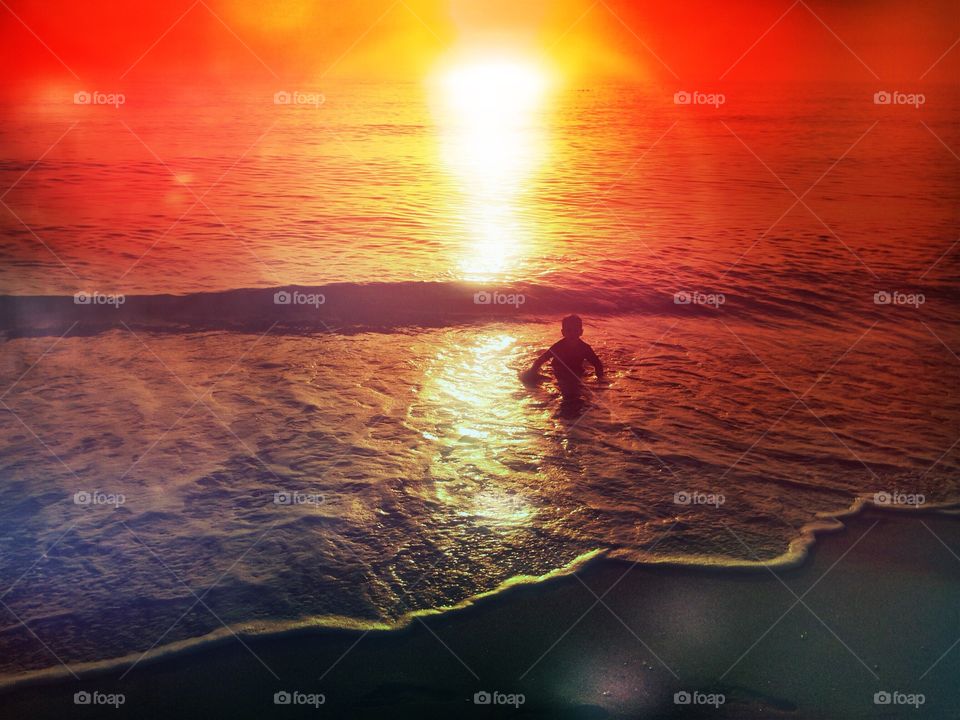 Small boy playing in the ocean surrounded by a dazzling red and gold sunset.