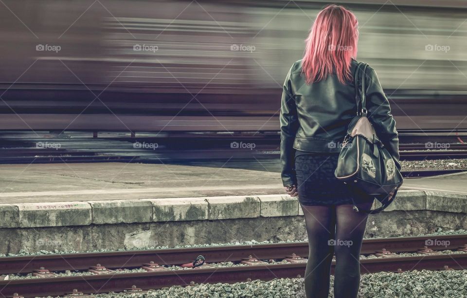 A woman in black with bright red hair stands on a platform as a rusty red, freight train whizzes past