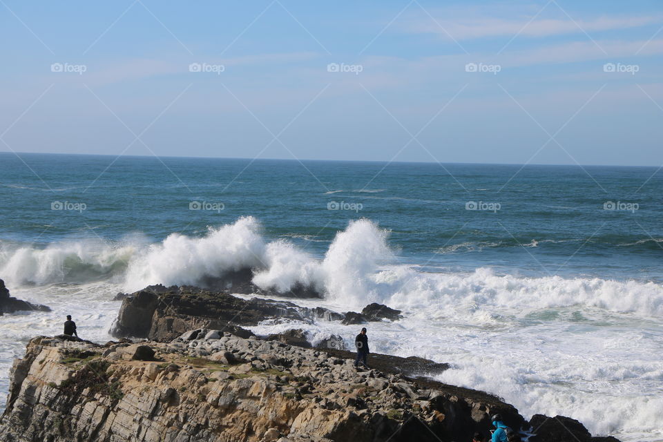 Blue Pacific with bib waves crashing on the big rocks by the shore while people walking on and watching the awesome scenery 