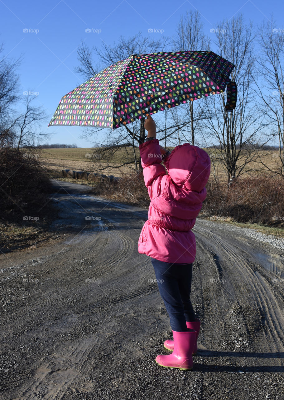 Little girl wearing pink rain boots and a pink coat putting up an umbrella