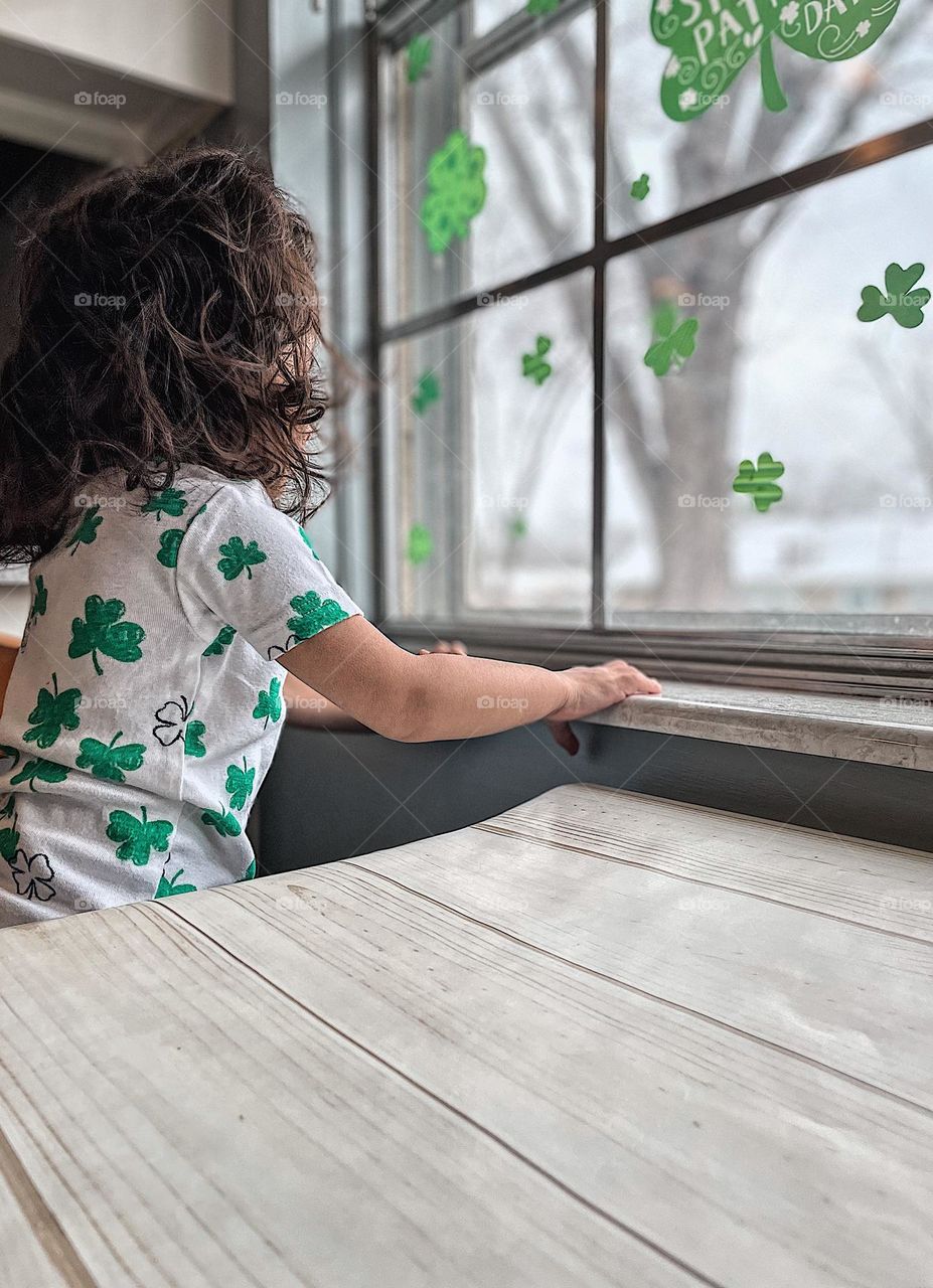 Toddler girl peers out window, toddler looks out window into backyard, toddler dressed in clover outfit looks out St. Patrick’s Day decorated windows, looking outside, little girl excited to go outside, Nosey neighbor toddler peers out window 