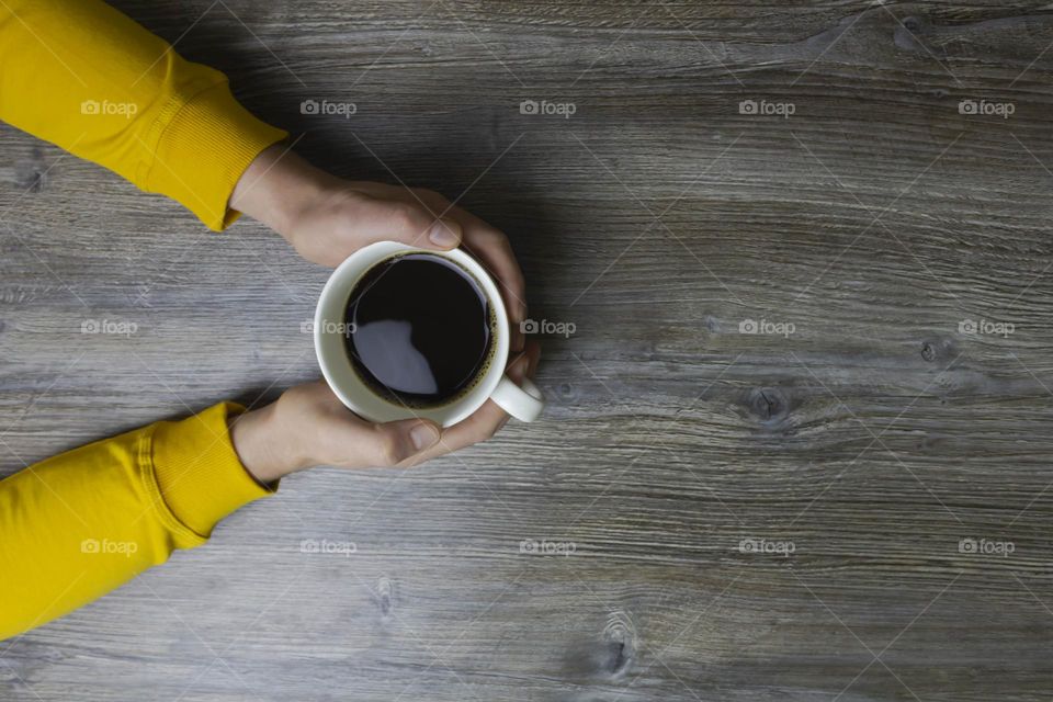 A man drinks coffee in the morning. Men's hands holding a white cup of coffee in a yellow jacket lie on a gray wooden surface, which is used as a background or a surface with incident light.