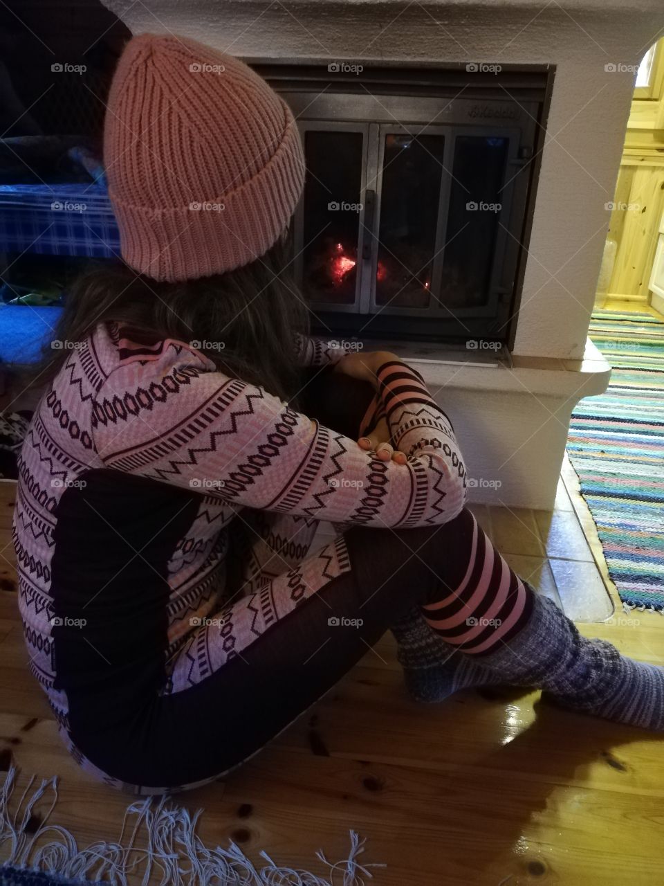 A young brunette woman sitting in the front of the fireplace on the varnished floor. She wears a knit cap, patterned underclothes and multicoloured striped woolen stockings, staring at the flames and ashes.