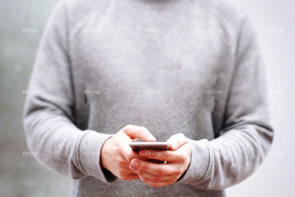 A mid section shot of a young man holding a mobile phone in his hands whilst texting and wearing neutral grey colours 