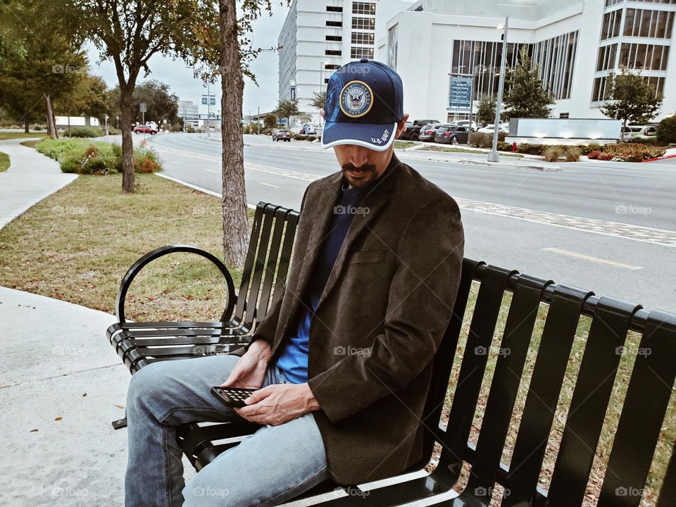 A man sitting on a city park bench on a fall day. He is wearing denim blue jeans a brown corduroy blazer and a blue shirt with a matching blue cap.