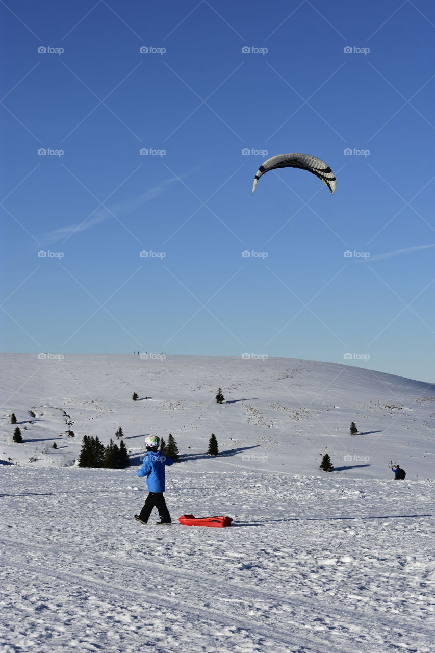 a little boy with his red sled in the snow and a kite in the blue sky