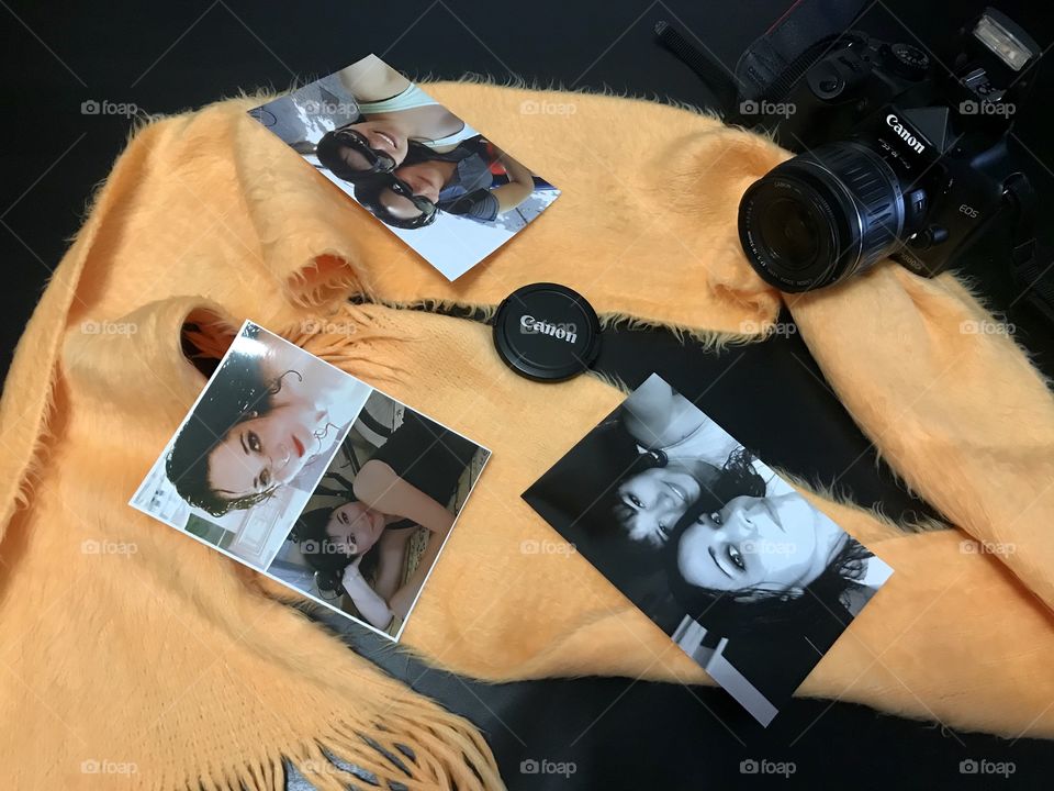 There is a Canon brand camera. There is a yellow scarf. On top of the scarf and around the camera there are photographs on paper.