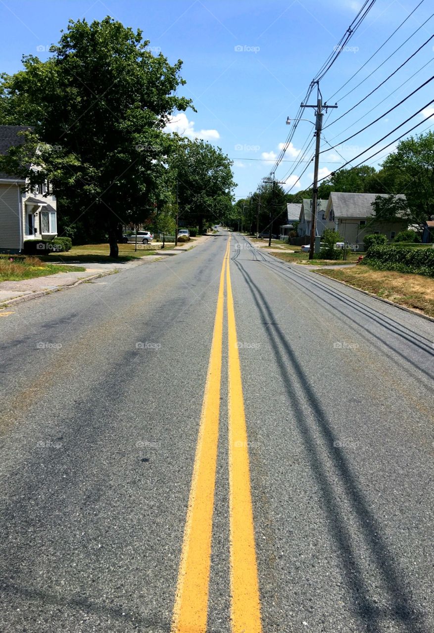 Backroads of America showing no cars on straight double yellow line street. Houses line both sides with one having a sidewalk.