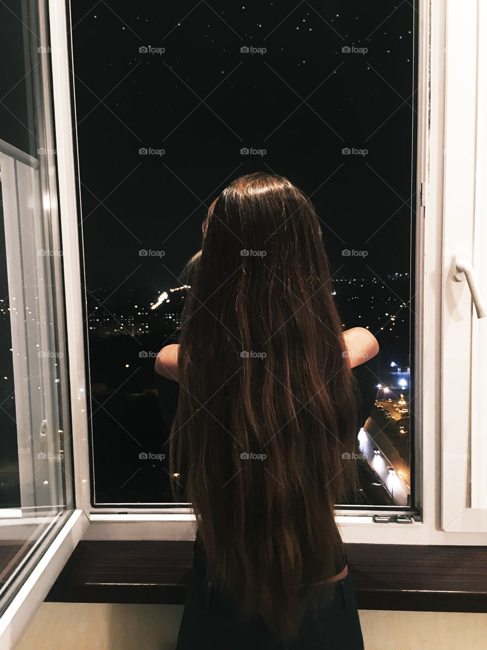 Young woman with long hair enjoying the night city lights view from the window 