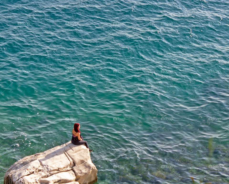 Summer, a young girl sits on the edge of a cliff and looks into the distance of a calm clear sea.  Summer vacation alone, relaxation
