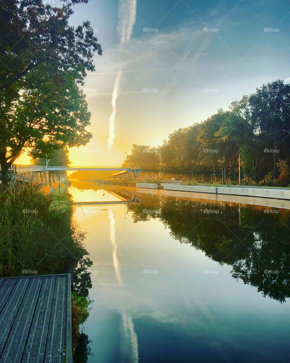 Gold glowing sunrise or sunset in a blue sky reflected in the water of a river or canal running through the forests