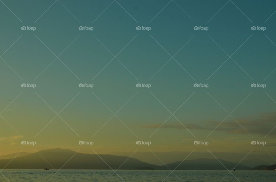 A clear bright sky showcasing a beautiful view of the bluish green and yellowish orange sunset on the beach with a view of the mountains at the back. 