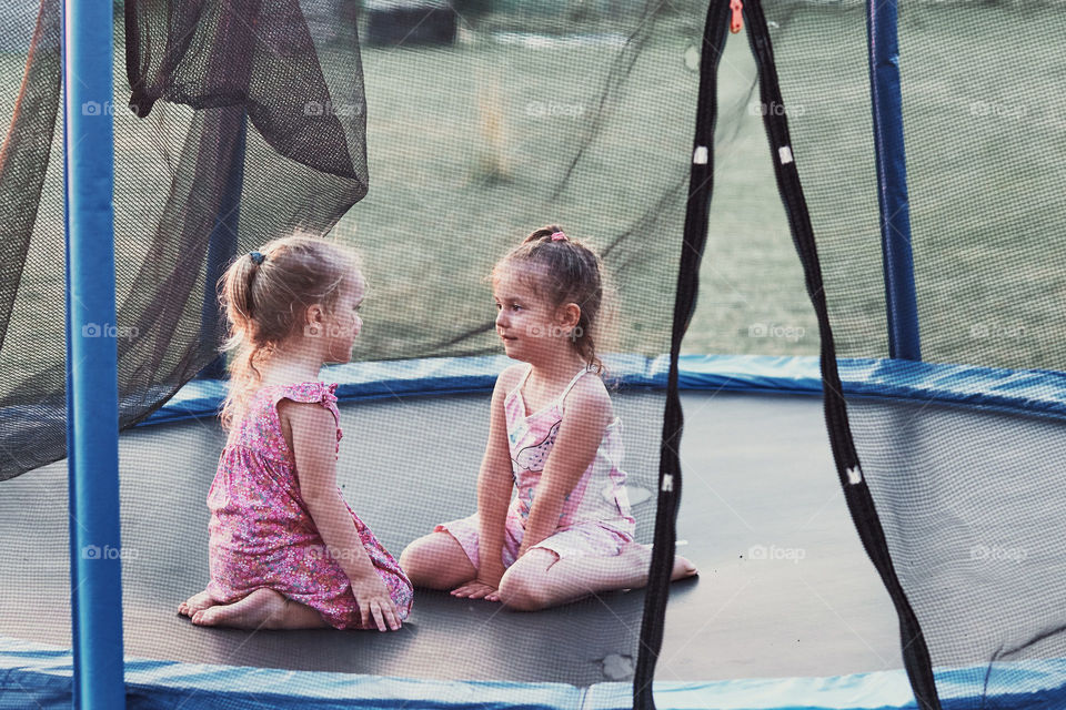 Two little cute girl talking, sitting and playing on trampoline in backyard on summer day. Candid people, real moments, authentic situations