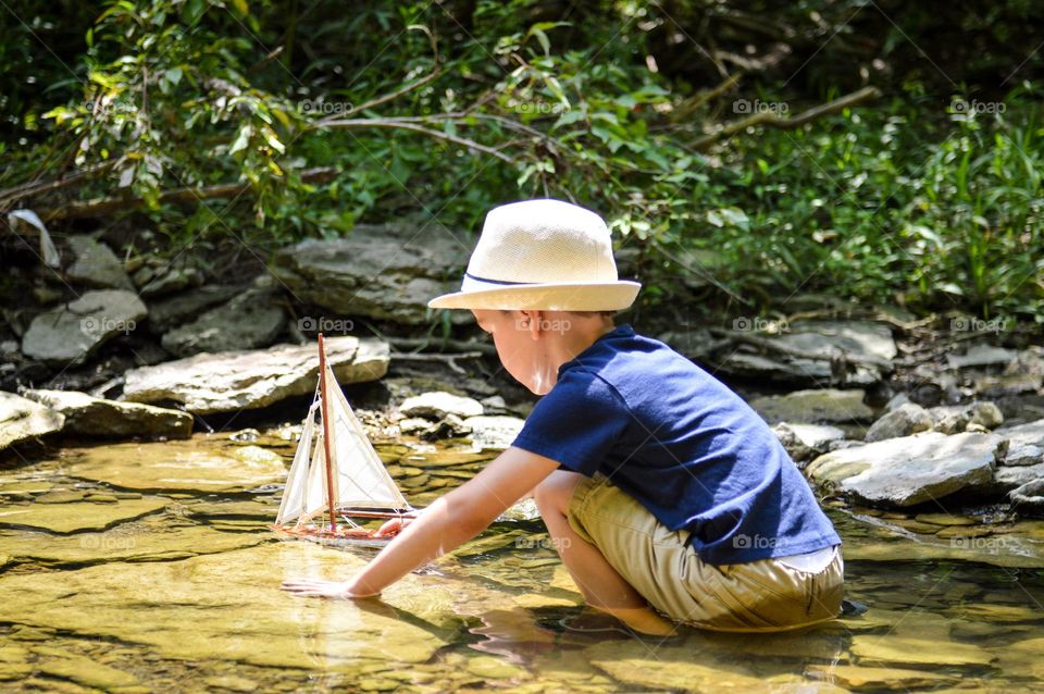 Young boy playing in a creek with a sailboat in the sunlight shining through the forest 