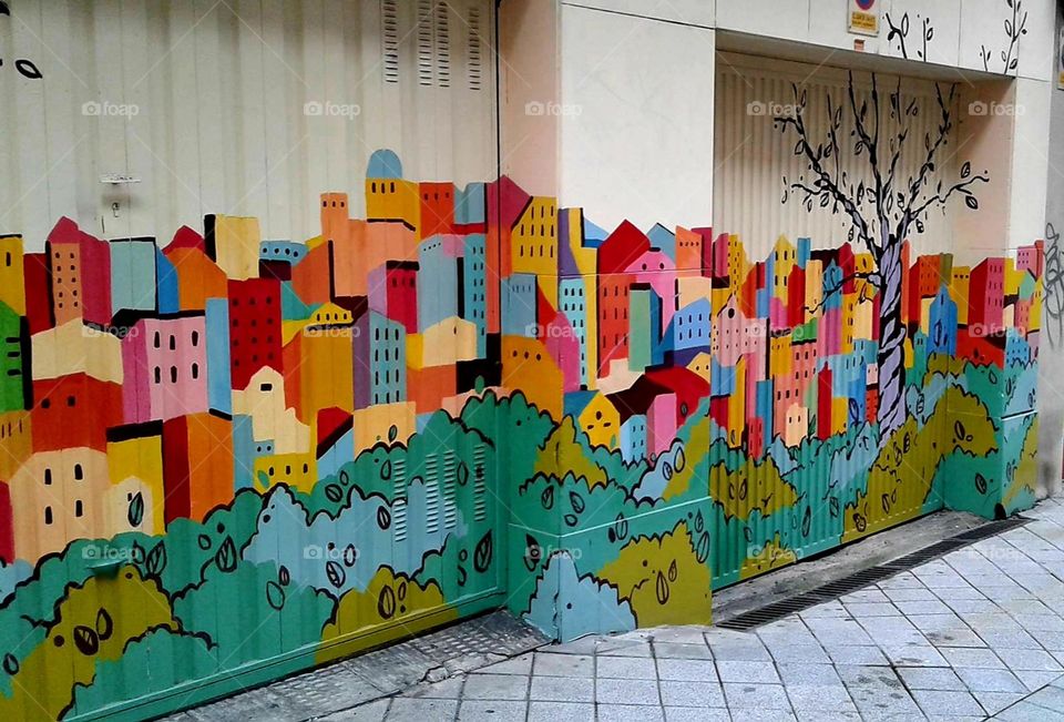 Visual Street Art: Painting of colorful city stretches down the side of a building along the sidewalk.