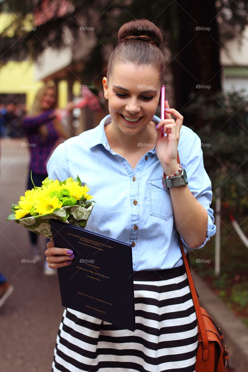 Happy woman walking on mobile phone holding flower bouquet and book 
