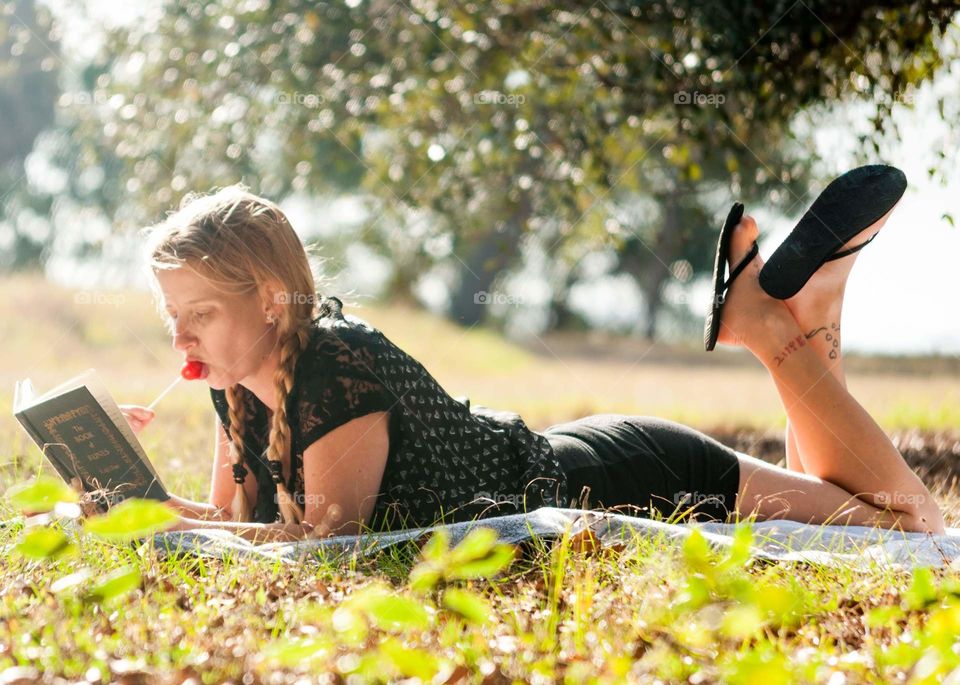 girl laying in the grass reading a book with a lollipop on a sunny day in the park