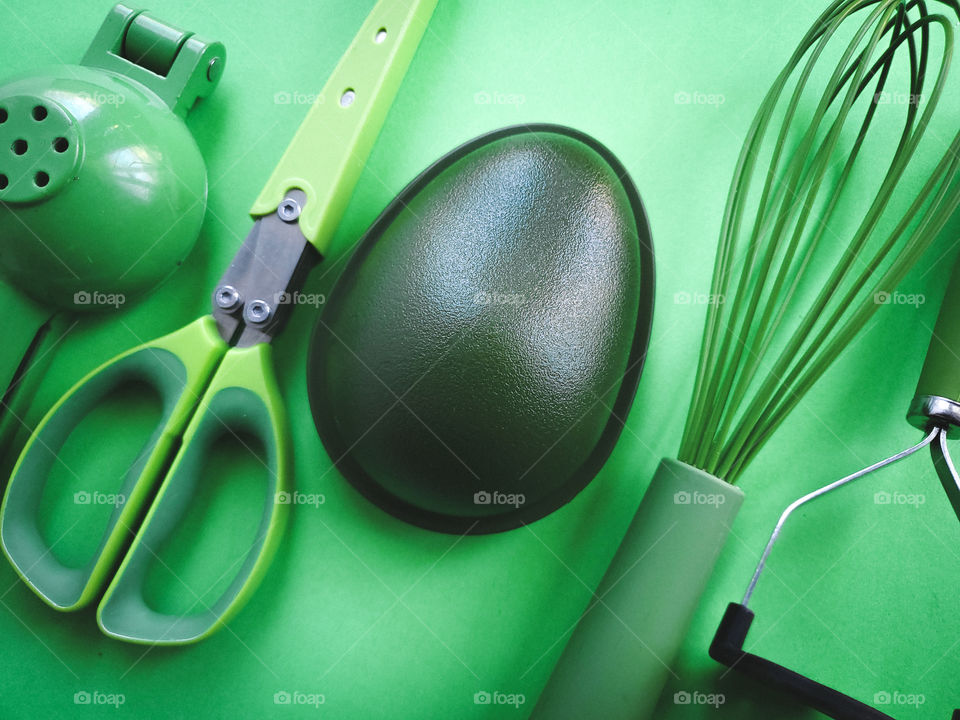 Five green kitchen utensils placed in a diagnol fashion: lemon/lime squeezer/press, herb mincing scissors,  avocado holder container, silicone coated wire wisk, and a masher.