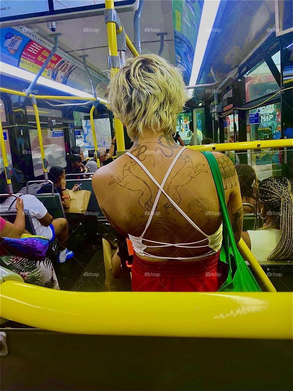 This picture of a lady riding the bus with bleached hair, a white crisscross top and bare back with tattoos is so streamlined in its angular momentum of the yellow metal tubing and neon lights it just has to be presented. 2023. Hypnotic Productions