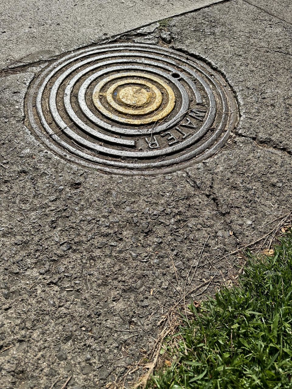 Round “WATER” concentric circle design manhole cover set in sidewalk near patch of green grass