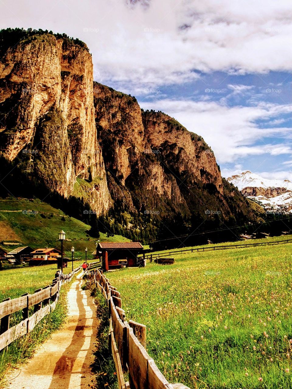 Landscape of the Dolomite mountains looking down a valley with a winding fenced path, large craggy golden hued mountains and snow capped mountains in the distance