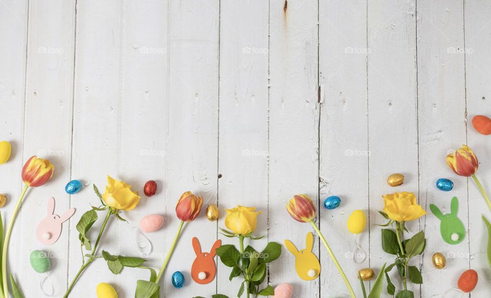 Yellow roses, fiery tulips, chocolate easter eggs, decorative eggs and wooden bunnies lie in a row from below on a white wooden table with copy space on top, flat lay close-up. Holiday banner concept